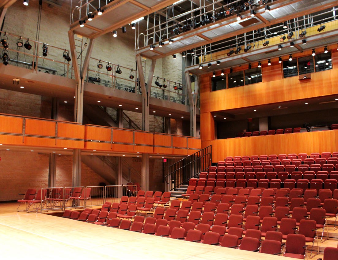 Modern theater auditorium with rows of red seats and overhead stage lighting.